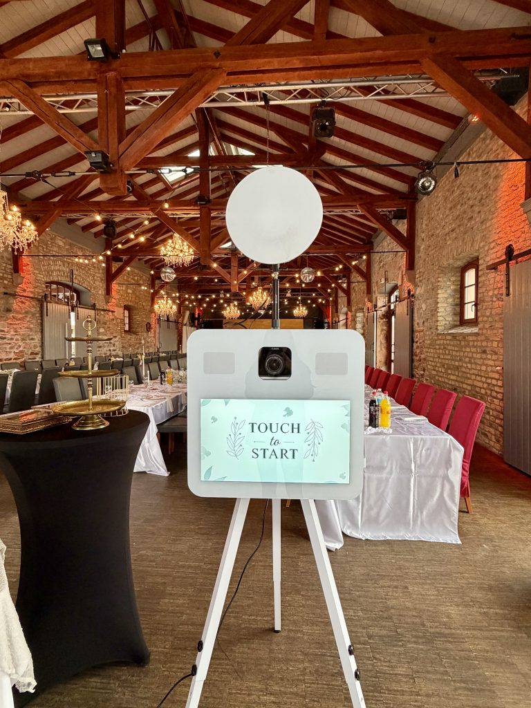 Weiße Fotobox mit Studioblitz in einer festlich gedeckten Scheune mit Holzbalken, Kronleuchtern und Lichterketten