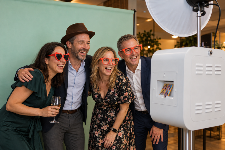 Vier Kollegen mit Fotoaccessoires neben einer weißen Fotobox vor einem mintfarbenen Hintergrund